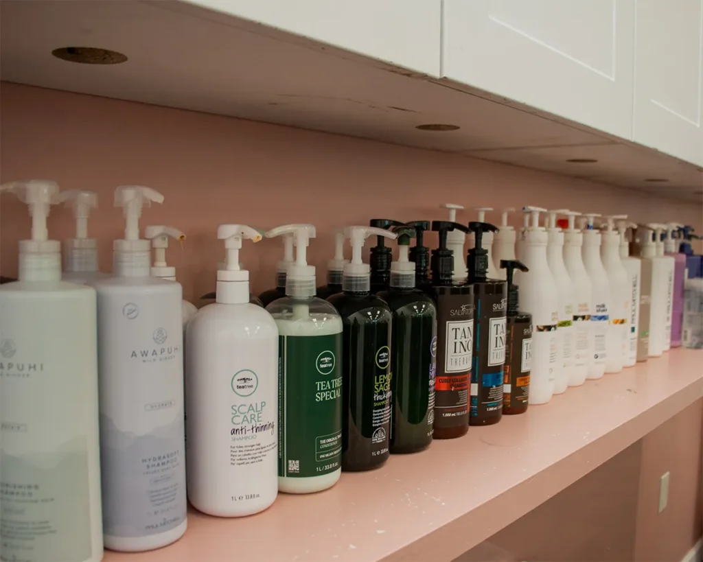 A row of assorted shampoo, conditioner, and hair treatment bottles with pump dispensers lined up on a shelf under a cabinet. - Salon Blissful, Mesa and Peoria, AZ