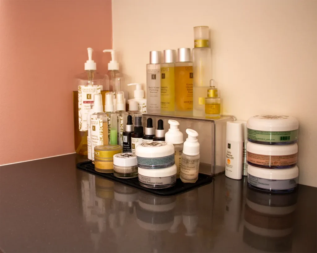 Various skincare and cosmetic products, including bottles, jars, and dropper vials, are neatly arranged on a bathroom countertop against a two-toned wall. - Salon Blissful, Mesa and Peoria, AZ