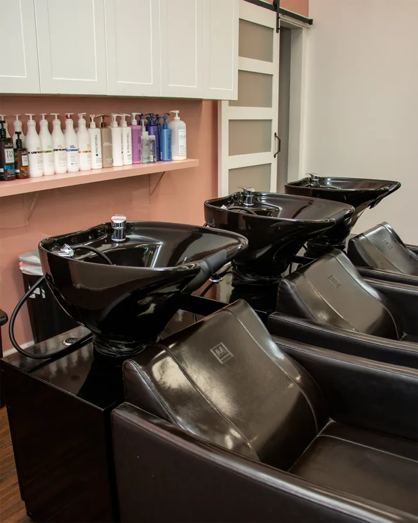 Three black salon shampoo stations with reclining chairs are positioned in front of a shelf holding various hair care products and bottles. - Salon Blissful, Mesa and Peoria, AZ
