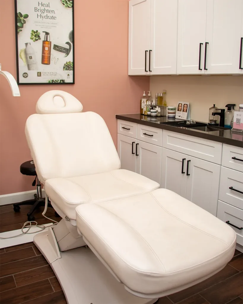 A clean treatment chair in a modern aesthetic clinic room with white cabinets, countertop products, and a skincare poster on a peach-colored wall. - Salon Blissful, Mesa and Peoria, AZ