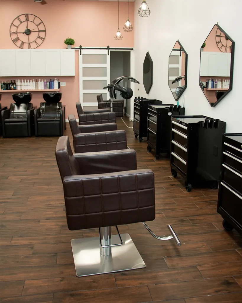 Modern hair salon with brown leather chairs, styling stations, hair-washing sinks, white cabinets, and wooden floors, with pendant lights and a large wall clock in the background. - Salon Blissful, Mesa and Peoria, AZ