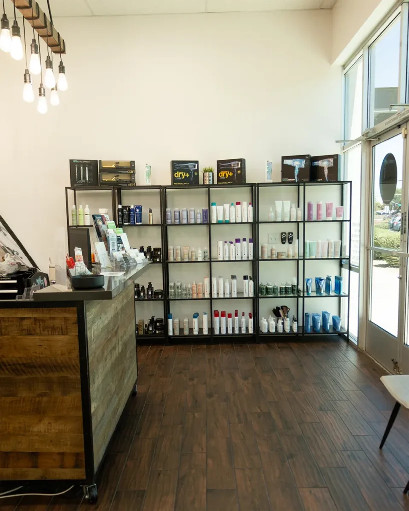 A modern salon retail area with shelves displaying various hair and beauty products beside a wooden reception desk and large windows. - Salon Blissful, Mesa and Peoria, AZ