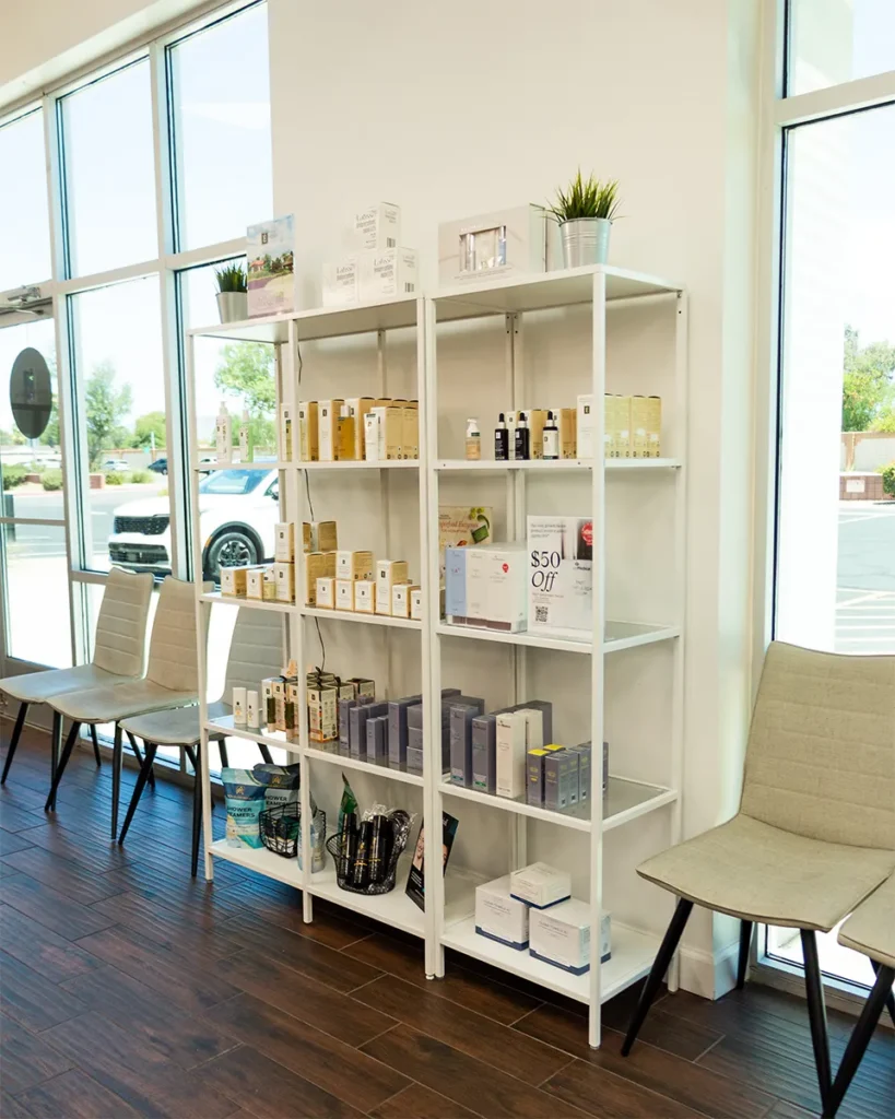 A white metal shelving unit displays various skincare and beauty products in a bright room with large windows and beige chairs. - Salon Blissful, Mesa and Peoria, AZ