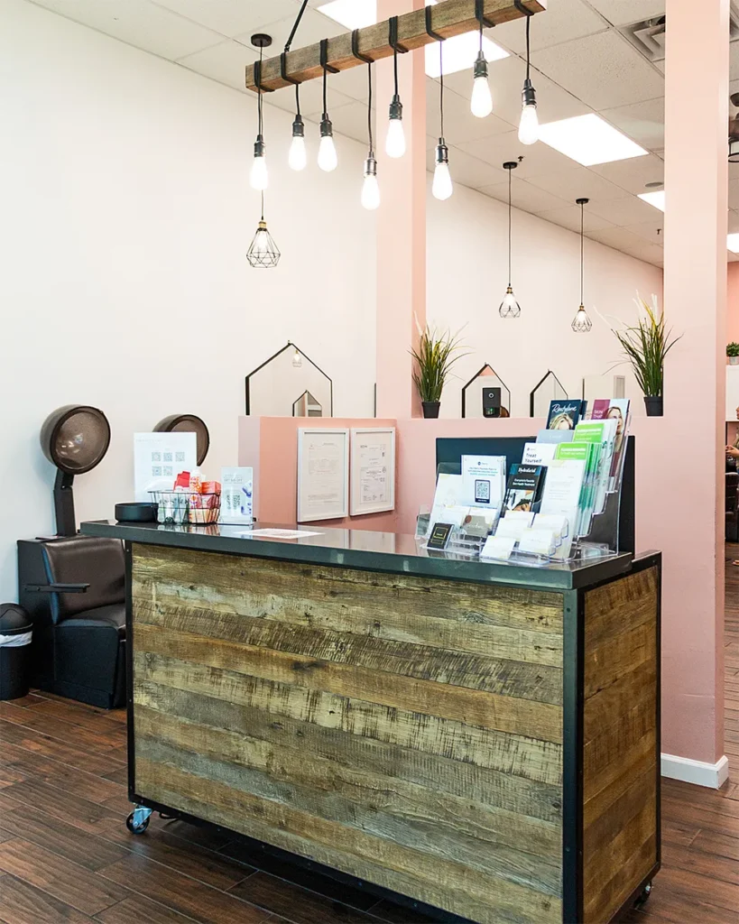 A salon reception area with a wooden front desk, brochures, and hanging light bulbs. Hair styling stations and hair dryers are visible in the background. - Salon Blissful, Mesa and Peoria, AZ